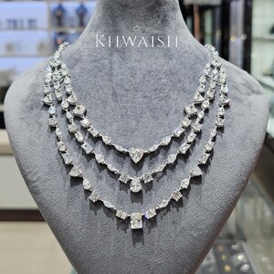 Puede incluir: Un collar de tres hilos en tono plateado con numerosas piedras transparentes facetadas en varias formas, incluyendo cortes de pera, esmeralda y redondos. El collar se exhibe sobre un busto de terciopelo gris y presenta el texto KHWAISH arriba.