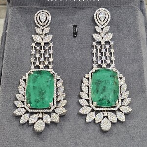 May include: A pair of long drop earrings featuring large, rectangular-cut green gemstones surrounded by pavé-set white stones. The design includes a leaf-like motif and a dangling lattice structure. The text KHWAISH is printed above the earrings.