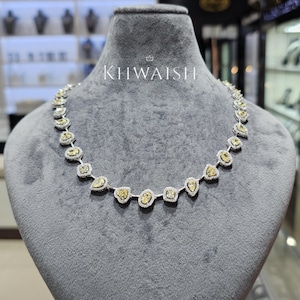 Puede incluir: Un collar que presenta una serie de diamantes amarillos en varias formas, incluyendo corte pera, ovalado y cojín, cada uno rodeado por un halo de pequeños diamantes blancos. La palabra KHWAISH está impresa en blanco sobre el collar en el expositor.