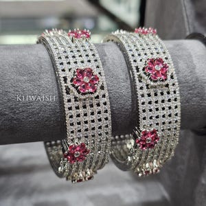 Pode incluir: Duas pulseiras de prata com um design de treliça, cravejadas com pequenas pedras transparentes. Cada pulseira apresenta múltiplos detalhes de flores rosa. O texto "KTIWAISH" é visível na pulseira esquerda.