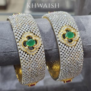 Puede incluir: Dos brazaletes plateados con un diseño floral, cada uno con una piedra preciosa verde central y rodeado de piedras claras más pequeñas. Los brazaletes tienen un interior dorado y se exhiben sobre una superficie gris. La marca "KHWAISH" es visible.