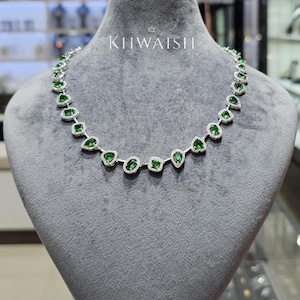 Puede incluir: Un collar de tono plateado que presenta una serie de piedras preciosas verdes alternadas en forma de pera y ovalada, cada una rodeada por un halo de pequeñas piedras blancas. El collar se exhibe en un busto de terciopelo gris con el texto KHWAISHI.