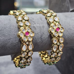 May include: Two gold bangles with a floral design. Each bangle features clear, round and teardrop-shaped stones, with a central pink stone in each floral cluster. The bangles are displayed on a grey surface.