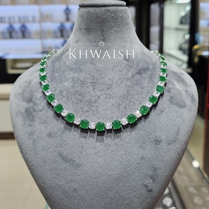 Puede incluir: Un collar plateado con piedras cuadradas verdes esmeralda y transparentes alternas. El collar se exhibe sobre un busto gris con el texto "KHWAISH" en blanco.
