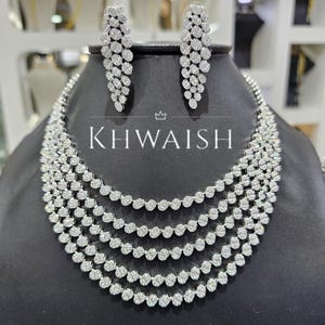 Può includere: Un set di gioielli abbagliante con una collana a più fili e orecchini abbinati. La collana ha più file di pietre scintillanti e gli orecchini hanno un design a cascata. Il testo "KHWAISH" è visualizzato sulla parte anteriore della collana.