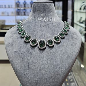 Puede incluir: Un collar que presenta una serie de piedras cabujón de color verde oscuro en forma ovalada, cada una rodeada por un halo de pequeñas piedras blancas transparentes. La pieza se exhibe sobre un busto de terciopelo gris, con el texto KHWAISH en blanco.