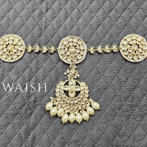 May include: Gold-toned headpiece with three circular, ornate sections connected by delicate chains. Each circle is embellished with clear stones and surrounded by small pearls. A central pendant with a floral design and dangling pearls adds elegance. The brand name "Khwaish" is visible.