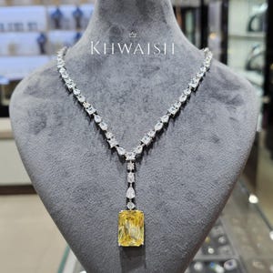 Puede incluir: Un collar de diamantes con un gran colgante rectangular amarillo. El collar presenta una serie de piedras claras y brillantes de varias formas. El collar se exhibe en una cabeza de maniquí gris. El texto "KHWAISH" es visible.