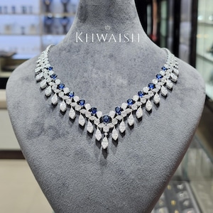 Puede incluir: Un collar de tono plateado con un diseño en forma de V, con zafiros azules en talla pera y numerosas piedras claras pequeñas en pavé. La pieza muestra el texto KHWAISH con un icono de corona encima, dispuesto en un patrón de hojas.