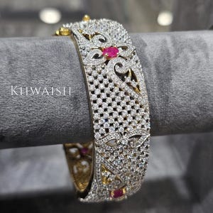 May include: A detailed, open-work bracelet with a gold-tone base, encrusted with numerous small, clear gemstones. The bracelet features floral and scroll designs, with small red gemstones as accents. The word "KUWAISH" is visible on the side.
