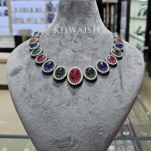 Könnte beinhalten: Eine Halskette mit ovalen, roten, blauen und grünen Cabochon-Edelsteinen, die jeweils von einem Kranz aus kleinen weißen Steinen in einer dunklen Metallfassung umgeben sind. Die Kette ist auf einer grauen Samtbüste ausgestellt.