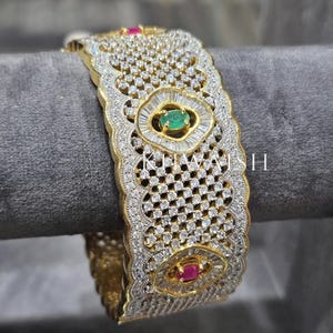 May include: A gold-coloured bracelet with detailed design. The bracelet has a repeating pattern of small, clear stones in a lattice design. Emerald and ruby-coloured stones are used as accents. The bracelet is displayed on a grey surface.