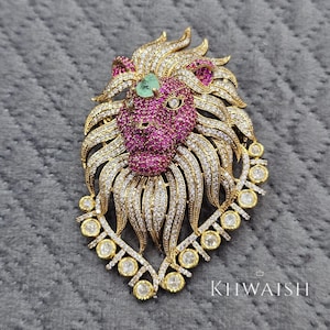 May include: A detailed lion-shaped pendant with a gold-tone frame, encrusted with sparkling white and pink gemstones. The lion's mane is adorned with white stones, and the face is a vibrant pink. An emerald-colored stone sits on the forehead. The pendant has the text "KHWAIISH".