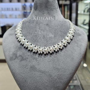 Puede incluir: Un collar de tono plateado con un patrón floral repetitivo compuesto por piedras transparentes brillantes y pequeñas perlas blancas. La pieza se exhibe sobre un busto de terciopelo gris e incluye el texto KHWAISH impreso en blanco arriba.