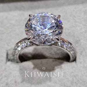 Puede incluir: Un deslumbrante anillo de compromiso con una gran gema redonda y transparente engastada en una banda plateada. La banda está incrustada con piedras más pequeñas y brillantes. El anillo se presenta en una caja gris. El texto "KIHWAISH" es visible.