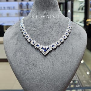 Puede incluir: Un collar en forma de V con una serie de piedras transparentes de corte marquesa y piedras azules redondas dispuestas en un patrón floral. La pieza se exhibe sobre un busto de terciopelo gris e incluye el texto KHWAISH impreso en blanco.
