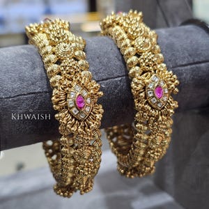 Op de afbeelding: Twee sierlijke gouden armbanden met ingewikkelde bloemen- en textuurontwerpen. Elke armband heeft een centrale ovale roze edelsteen omringd door heldere stenen. De tekst "KHWAISH" is zichtbaar op de linkerarmband.
