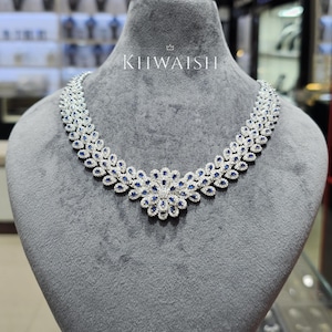 Puede incluir: Un collar de tono plateado con un motivo floral central y eslabones simétricos en forma de hoja. La pieza está incrustada con numerosas piedras claras pequeñas y piedras azul oscuro en forma de lágrima. El texto KHWAISH aparece sobre el collar.