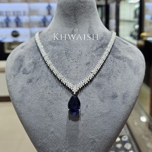 Puede incluir: Un collar de diamantes con un colgante en forma de pera de color azul oscuro. El collar se exhibe en un maniquí gris. El texto "KHWAISH" es visible.