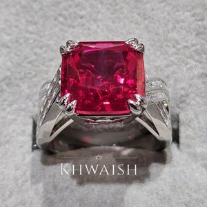 Puede incluir: Un anillo plateado con una gran gema roja vibrante de forma rectangular. La banda del anillo está detallada con piedras más pequeñas y claras y un intrincado trabajo en metal. El texto "KHWAISH" es visible en la base del anillo.