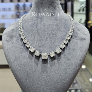 Puede incluir: Un collar de tono plateado con una serie de piedras transparentes de corte cuadrado dispuestas en línea graduada, con una piedra central más grande. La pieza se exhibe sobre un busto de terciopelo gris e incluye el texto KHWAISH en blanco.