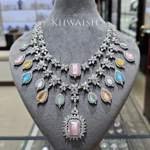 May include: A dazzling silver necklace with a floral design, featuring a large pink rectangular center stone and multiple teardrop-shaped gemstones in various pastel colors. The necklace is displayed on a gray mannequin with the text "KHWAISH".