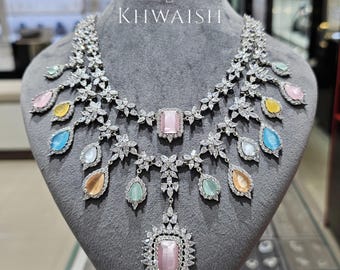 May include: A dazzling silver necklace with a floral design, featuring a large pink rectangular center stone and multiple teardrop-shaped gemstones in various pastel colors. The necklace is displayed on a gray mannequin with the text "KHWAISH".