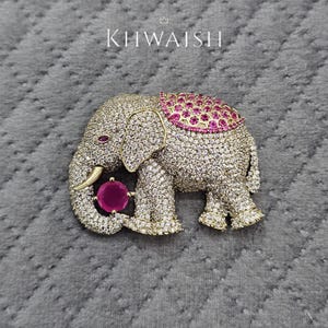 Puede incluir: Broche de elefante dorado incrustado con cristales transparentes, con una piedra de color rubí en la trompa y en la espalda. El broche tiene la palabra "KHWAISH" en blanco en la parte superior.