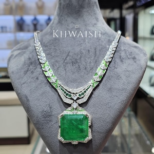 May include: A luxurious necklace with a large, emerald-cut green gemstone pendant. The necklace features a silver-colored chain with green and white accents. The brand name "KHWAISH" is displayed above the necklace on a gray mannequin.