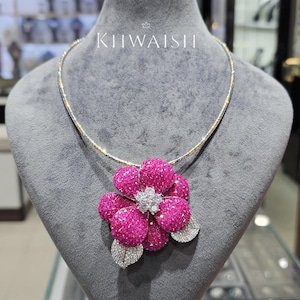 Op de afbeelding: Een ketting met een grote bloemvormige hanger bezet met levendige roze stenen, een centrum van heldere witte stenen en twee zilverkleurige bladeren ingelegd met heldere steentjes. De tekst KIIWAISHI staat boven de bloemvormige hanger.