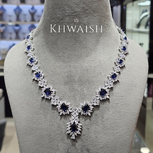 Puede incluir: Un collar de tono plateado que presenta una serie de piedras de corte rectangular azul profundo alternadas con grupos de piedras claras de corte marquesa. El centro muestra un colgante de gota prominente. El texto KHWAISH aparece impreso en blanco.