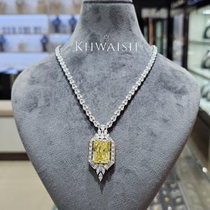 May include: A diamond necklace featuring a large, rectangular yellow gemstone pendant. The pendant is framed by smaller diamonds and suspended from a delicate chain. The necklace is displayed on a grey mannequin. The text "KHWAISH" is visible.