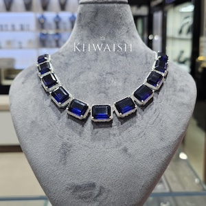 Puede incluir: Un collar que presenta una serie de piedras rectangulares de color azul intenso, cada una rodeada por un borde de pequeñas piedras claras y brillantes. La pieza se exhibe sobre un busto de terciopelo gris, con el texto KHWAISH impreso en blanco.