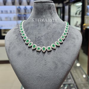 Puede incluir: Un collar que presenta una serie de piedras preciosas verdes alternadas en forma de pera y corte cuadrado, cada una rodeada por un halo de pequeñas piedras blancas engastadas en metal plateado. La palabra KHWAISH aparece en blanco sobre el centro.