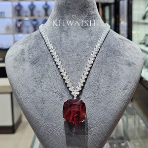 May include: A silver-toned necklace featuring a large, square-cut deep red gemstone pendant suspended from a chain composed of numerous small, clear, sparkling stones. The word KHWAISH is printed in white above the necklace on the display.