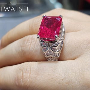 May include: A silver-toned ring featuring a large, rectangular, vibrant pink center stone held by four prongs. The band is encrusted with multiple rows of clear, baguette-cut stones. The text KHWAISH is visible in the upper left corner of the image.