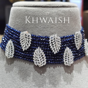 Può includere: Una collana girocollo blu navy con abbellimenti argentati a forma di foglia. La collana presenta più fili di piccole perline blu scuro ed è adornata da sei ciondoli a forma di foglia incastonati con piccole pietre scintillanti. La parola "KHWAISH" è visualizzata.