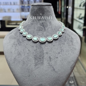 May include: A silver-toned necklace featuring a series of oval-shaped, light mint green stones. Each stone is surrounded by a halo of small, clear, sparkling crystals. The brand name KHWAISH is printed in white text above the necklace.