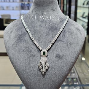 May include: A silver-toned necklace featuring a central oval-shaped dark green gemstone surrounded by clear white stones. The pendant has a cascading fringe design with multiple teardrop-shaped stones. The text KHWAISH is printed above the necklace.
