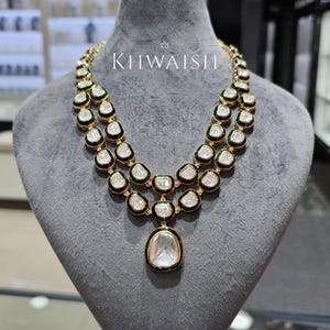 Puede incluir: Un collar dorado con dos hileras de piedras preciosas transparentes engarzadas. Las piedras están delineadas en negro y verde, con un gran colgante en forma de pera en la parte inferior. El collar se exhibe en un maniquí gris. El texto "KIIWAISHI" es visible.