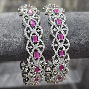 May include: Two silver-toned bangles with a repeating pattern of pink rectangular stones and clear, round accent stones. The bangles have an openwork design and are displayed on a gray surface. The text "KIIWAISHI" is visible on the left bangle.