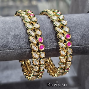 Könnte beinhalten: Zwei goldene Armreifen mit abwechselnd klaren und rosa Edelsteinen. Die Armreifen haben ein detailliertes Design mit einem grünen Rand. Der Markenname "KHWAISH" ist unten sichtbar.
