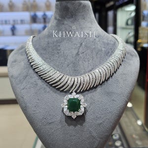 Może przedstawiać: Olśniewający srebrny naszyjnik z dużym wisiorkiem z zielonym kamieniem szlachetnym o szlifie szmaragdowym. Naszyjnik posiada szereg małych, błyszczących kamieni ułożonych w zakrzywiony wzór. Nazwa marki "KHWAISH" jest wyświetlana białym tekstem.