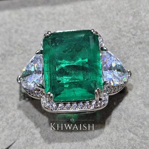 Puede incluir: Un anillo plateado con una gran piedra verde rectangular con talla esmeralda como pieza central. Dos piedras claras en forma de pera flanquean la esmeralda. El anillo está engastado con piedras claras más pequeñas alrededor de la esmeralda y a lo largo de la banda. La palabra "KHWAISH" es visible.