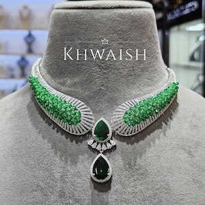 Könnte beinhalten: Eine luxuriöse Halskette mit dem Wort "KHWAISH" in weißen Großbuchstaben. Die Halskette hat ein silberfarbenes, geschwungenes Design mit grünen Edelsteinen und einem großen, tropfenförmigen grünen Edelsteinanhänger.