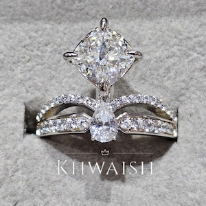 Peut inclure: Un ensemble de bagues de fiançailles en diamant avec une grosse pierre centrale de taille carrée et une pierre d'accent en forme de poire. La bague est ornée de petits diamants. Le texte "KIIWAISH" est visible.