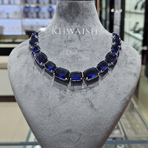Puede incluir: Un collar que presenta una serie de grandes piedras rectangulares de color azul intenso con facetas, engastadas en metal de tono plateado. Las piedras están dispuestas en una sola fila y el texto KIIWAISHI está impreso en blanco sobre el centro.