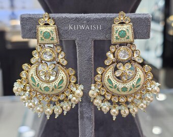 May include: A pair of ornate gold-toned earrings with a cream and green enamel design. The earrings feature a square top, a large central stone, and a crescent-shaped bottom with pearl and bead accents. The text "KHIWAISH" is visible.