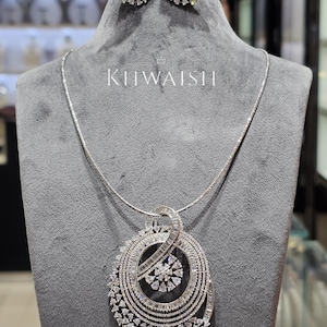 May include: A silver-toned jewelry set featuring a pendant necklace and matching earrings. The necklace has a large, circular pendant with a floral design and is paired with round earrings. The brand name "KIIWAISHI" is displayed on the gray mannequin.