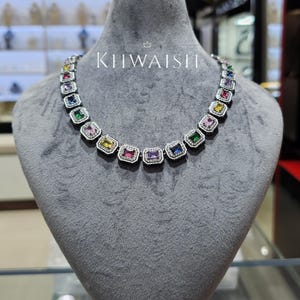 Puede incluir: Un collar plateado con una serie de piedras preciosas de colores, de corte cuadrado. Cada gema está enmarcada por un halo de piedras más pequeñas y transparentes. El collar se exhibe en un maniquí gris con el texto "KHWAISH" encima.